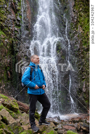 Tourist attraction of Germany - falls of Burgbach Waterfall near Schapbach, Black Forest, Baden-Wurttemberg, Germany. Man hiker in blue jacket standing on stone and looks at flow of falling water Tourist attraction of Germany - falls of Burgbach Waterfall near Schapbach, Black Forest, Baden-Wurttemberg, Germany. Man hiker in blue jacket standing on stone and looks at flow of falling water 104242967