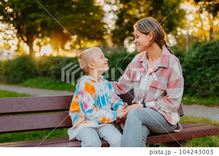mother and son sit on a park bench in the rays of the setting sun. the concept of a family. Mother's Day. beautiful girl (mother) with a boy (son) in the park in the park are sitting on a bench at mother and son sit on a park bench in the rays of the setting sun. the concept of a family. Mother's Day. beautiful girl (mother) with a boy (son) in the park in the park are sitting on a bench at 104243003