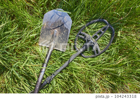 A metal detector and a shovel for searching for coins and precious metal products lie crossed on the green grass 104243318