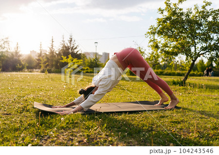 Side view of healthy female yoga practitioner preforming Downward Facing Dog Pose Adho Mukha Svansana at city park on sunny summer day. Concept of outdoors yoga, fitness, active and sporty lifestyle. 104243546