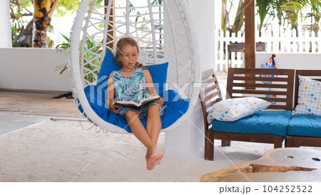Little girl reading book on tropical terrace, resting in hanging swing chair Little girl reading book on tropical terrace, resting in hanging swing chair 104252522