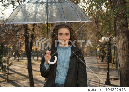 Portrait of cute woman with long hair under transparent umbrella. Beautiful girl stands on alley of fall park 104252948
