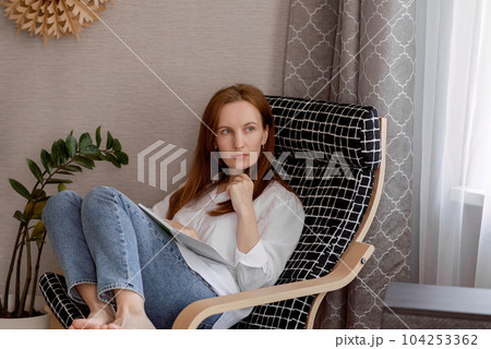 A young smiling woman, a happy confident woman in her 30's, holding a BLOCKET OR BOOK, sitting in a chair at home, working, looking at the camera. High quality photo 104253362
