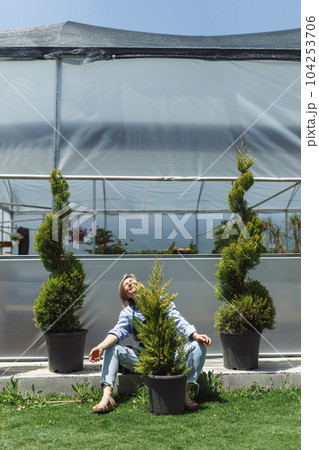 Portrait of smiling young botanist holding a fresh flower plant. Young woman holding small tree in pot in gardening center. Successful botanist and greenhouse owner concept Portrait of smiling young botanist holding a fresh flower plant. Young woman holding small tree in pot in gardening center. Successful botanist and greenhouse owner concept 104253706
