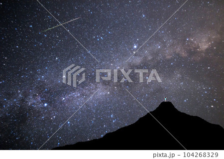 天の川と流れ星を山頂でガッツボーズしながら見つける男性。夏の天体観測イメージ。  104268329