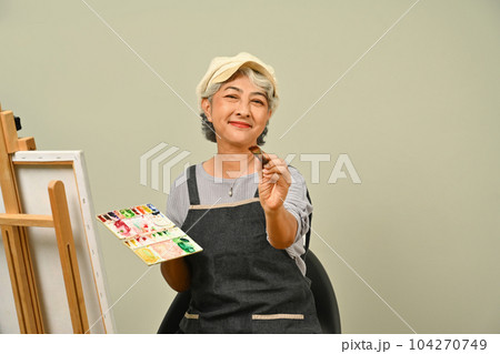 Beautiful 60s senior woman wearing apron holding brush and palette over gray background Beautiful 60s senior woman wearing apron holding brush and palette over gray background 104270749