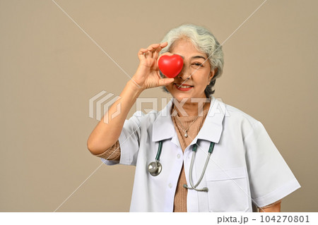 Gray haired 60s female cardiologist holding and pointing at red heart. Cardiology, medicine, donation and healthcare concept Gray haired 60s female cardiologist holding and pointing at red heart. Cardiology, medicine, donation and healthcare concept 104270801
