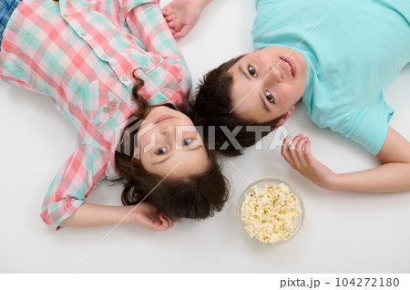 View from above of Caucasian adorable kids, beautiful little girl in checkered shirt and handsome preteen boy in blue t-shirt, lying on white background with a bowl of popcorn, looking at camera 104272180