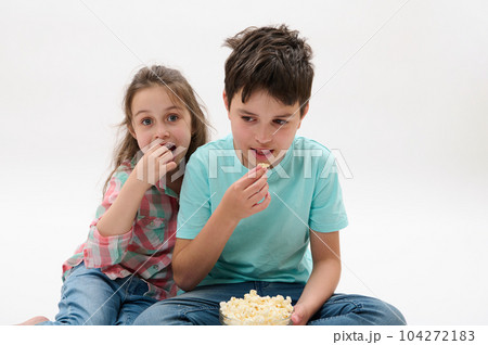 Caucasian adorable cheerful happy kids, preteen boy and little girl eating popcorn, watching movie or cartoons, smiling looking toward the camera, sitting together on isolated white studio background 104272183
