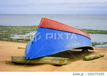 A blue-red boat lies upside down on the sandy seashore. 104275260