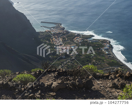 Aerial view of village resort Vueltas with port, Seen from top of La Merica mountain cliffs, Camino La Merica hiking trail. Valle Gran Rey, La Gomera, Canary Islands. Copy space. Aerial view of village resort Vueltas with port, Seen from top of La Merica mountain cliffs, Camino La Merica hiking trail. Valle Gran Rey, La Gomera, Canary Islands. Copy space. 104277756