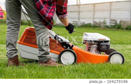 Unrecognisable gardener man in protective gloves starts the lawnmower before cutting green grass lawn in his backyard. Man with motorised lawnmower cares for landscaping lawn Unrecognisable gardener man in protective gloves starts the lawnmower before cutting green grass lawn in his backyard. Man with motorised lawnmower cares for landscaping lawn 104279196