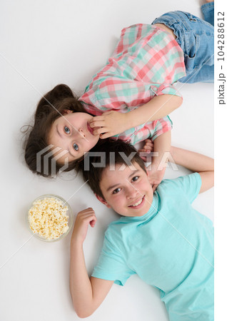 Top view happy kids, pre teen boy and little girl, brother and sister smiling looking at camera, lying on their backs on white studio background near a bowl with tasty popcorn, expressing positivity Top view happy kids, pre teen boy and little girl, brother and sister smiling looking at camera, lying on their backs on white studio background near a bowl with tasty popcorn, expressing positivity 104288612
