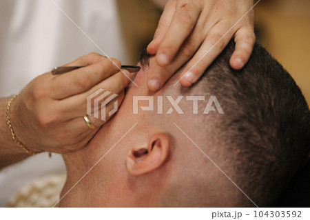 Close-up of barber man trimming eyebrows with tweezers for confident man Close-up of barber man trimming eyebrows with tweezers for confident man 104303592