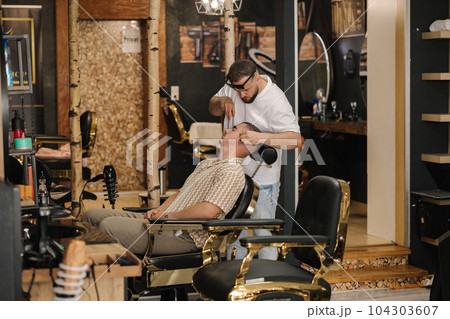 Side view process of threading procedure in barber shop. Professional barber correcting shape of brows with threads male client sitting in chair. Side view process of threading procedure in barber shop. Professional barber correcting shape of brows with threads male client sitting in chair. 104303607