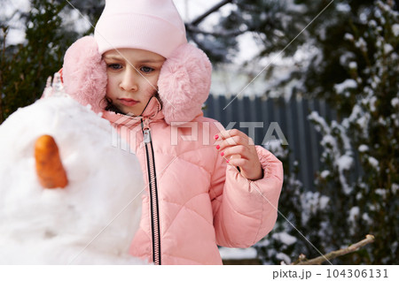 Caucasian adorable child 6 years old, lovely little girl in pink warm winter clothes and fluffy ear muffs, building a snowman in the snow covered backyard. Winter leisures activity. Christmas time Caucasian adorable child 6 years old, lovely little girl in pink warm winter clothes and fluffy ear muffs, building a snowman in the snow covered backyard. Winter leisures activity. Christmas time 104306131