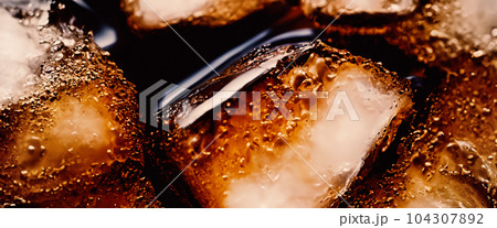 Cola with Ice. Close up of the ice cubes in cola water. Texture of carbonate drink with bubbles in glass. Cola soda and ice splashing fizzing or floating up to top of surface. Cold drink background. Cola with Ice. Close up of the ice cubes in cola water. Texture of carbonate drink with bubbles in glass. Cola soda and ice splashing fizzing or floating up to top of surface. Cold drink background. 104307892