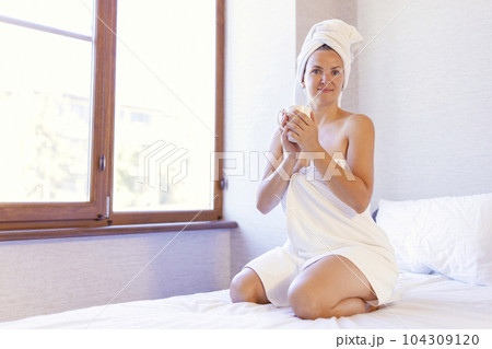 Woman in bath towel and towel on hair holding a cup and smiling 104309120