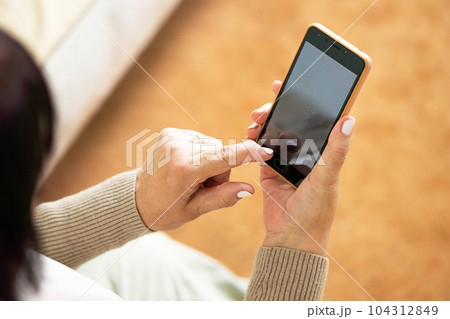 Close up of hands from middle aged woman sitting on sofa at home and using a mobile phone. Close up of hands from middle aged woman sitting on sofa at home and using a mobile phone. 104312849