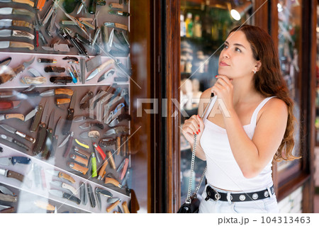 Interested young woman viewing various knifes placed on display case Interested young woman viewing various knifes placed on display case 104313463