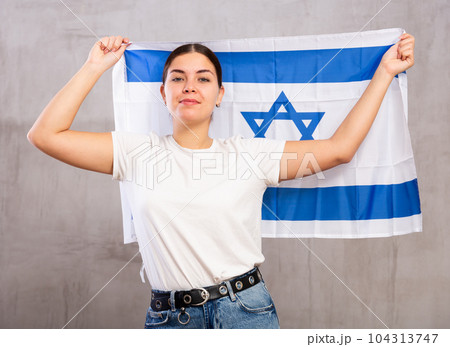 Portrait of serious young female holding national flag of Israel against gray wall Portrait of serious young female holding national flag of Israel against gray wall 104313747