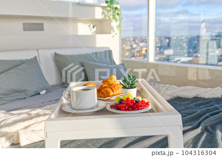 White Tray with tasty breakfast on bed in light bedroom with big windows in skyscraper. lifestyle White Tray with tasty breakfast on bed in light bedroom with big windows in skyscraper. lifestyle 104316304