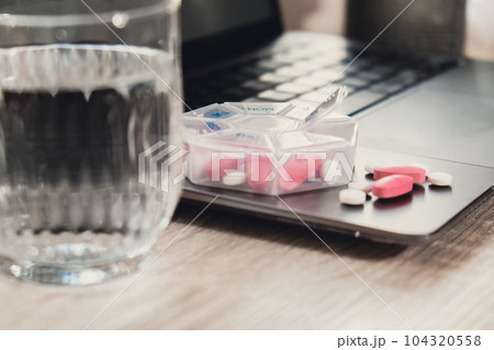 Organizer weekly shots with glass of water on workplace background and laptop Closeup of medical pill box with doses of tablets for daily take medicine with white pink drugs and capsules. Health care Organizer weekly shots with glass of water on workplace background and laptop Closeup of medical pill box with doses of tablets for daily take medicine with white pink drugs and capsules. Health care 104320558