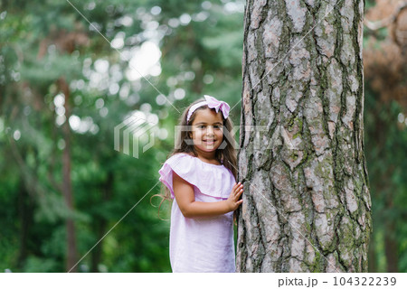 A beautiful girl in a pink dress stands near a tree in the forest in summer, smiling and dreaming 104322239