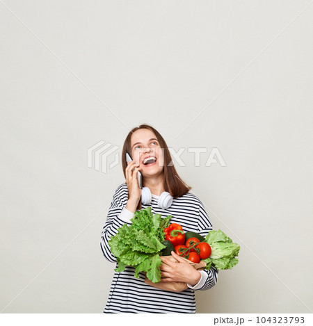 Amazed woman with brown hair wearing striped shirt standing isolated over gray background talking on mobile phone looking up at advertisement area holding vegetables 104323793