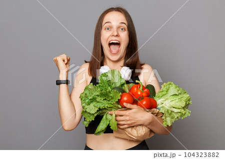 Overjoyed happy Caucasian woman holding fresh organic vegetables standing isolated over gray background clenched fist buying food in shop or market screaming with happiness. Overjoyed happy Caucasian woman holding fresh organic vegetables standing isolated over gray background clenched fist buying food in shop or market screaming with happiness. 104323882