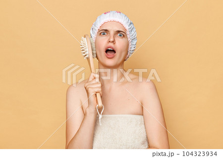 Shocked scared caucasian woman with massage brush on beige background taking shower, looking at camera with open mouth and displeased expression. 104323934