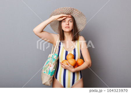 Shocked surprised astonished woman wearing striped swimsuit and straw hat isolated gray background standing with fresh oranges keeps hand near forehead looking far. Shocked surprised astonished woman wearing striped swimsuit and straw hat isolated gray background standing with fresh oranges keeps hand near forehead looking far. 104323970