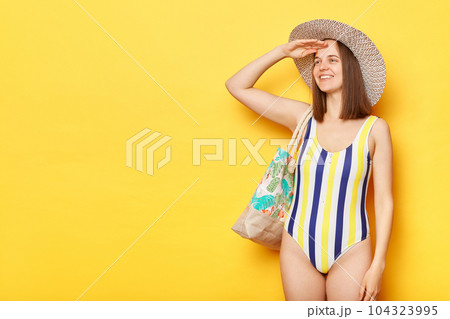 Cute smiling woman wearing swimsuit isolated on yellow background looking far with hand near forehead expressing positive emotions copy space for promotions. Cute smiling woman wearing swimsuit isolated on yellow background looking far with hand near forehead expressing positive emotions copy space for promotions. 104323995