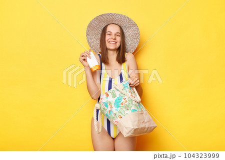 Satisfied woman wearing swimsuit isolated on yellow background standing with beach bag in straw hat holding her protective spf cream enjoying of quality. 104323999