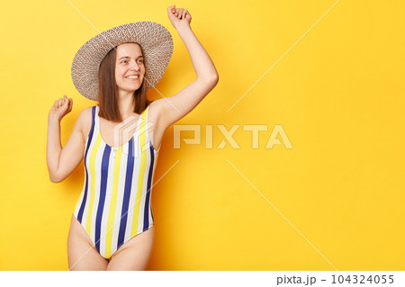 happy overjoyed woman wearing striped swimming suit and hat isolated on yellow background dancing rejoicing her vacation copy space for promotional text. 104324055