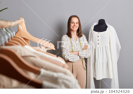 Confident successful female sewer standing near mannequin with new dress over gray background posing in her atelier looking at camera folded hands. 104324320