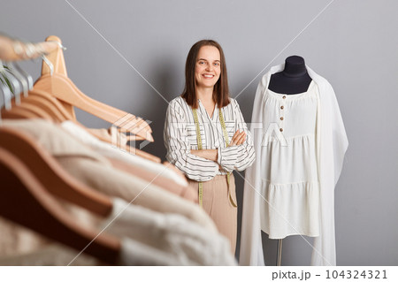 Unique garments, happy customers. Attractive smiling woman tailor standing near new dress on mannequin iisolated over gray background keeps hands crossed looking at camera. 104324321