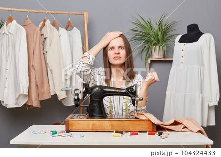 Sad upset woman sewer posing at her workplace sewing clothing, showing facepalm gesture, made mistake, dressmaker making wrong stitch, spoiled material for dress. Sad upset woman sewer posing at her workplace sewing clothing, showing facepalm gesture, made mistake, dressmaker making wrong stitch, spoiled material for dress. 104324433