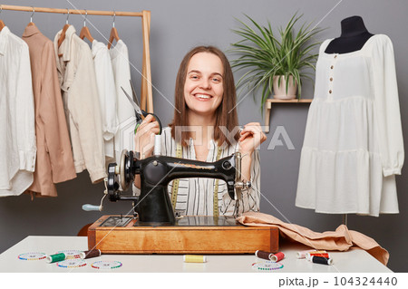 Cheerful caucasian female dressmaker businesswoman working on sewing machine in her own tailor workshop, holding scissors looking at camera with positive facial expression. Cheerful caucasian female dressmaker businesswoman working on sewing machine in her own tailor workshop, holding scissors looking at camera with positive facial expression. 104324440