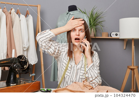 Shocked caucasian young adult female sewer working in atelier, talking on mobile phone, making facepalm gesture, having problems with supply of new fabrics. Shocked caucasian young adult female sewer working in atelier, talking on mobile phone, making facepalm gesture, having problems with supply of new fabrics. 104324470