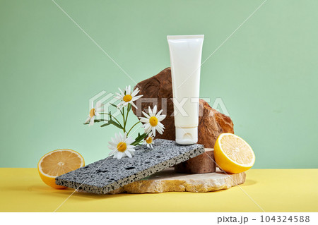 An empty white plastic tube placed on brick and stone with fresh half of lemon and flower on a pastel background. Blank label for branding mockup, natural vitamin C skincare beauty product concept. 104324588