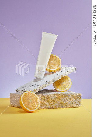 On a pastel purple background, white plastic tube displayed with marble brick, block of stone and halves of lemon. Cosmetic mockup for advertising and branding with lemon extract. Front view 104324589