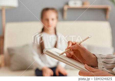 Closeup of doctor psychologist holding paper notebook making notes while talking with small girl at psychological therapy session meeting, blur defocused little kid patient. 104324825