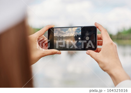 Unrecognizable female blogger using cellular technology for shooting video content during travel vacations, woman with smartphone device photographing lake or river. 104325016