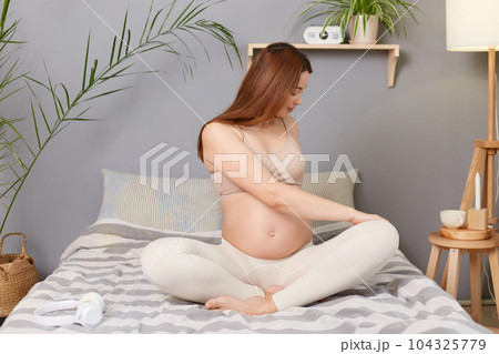 Attractive pregnant brown haired caucasian woman sitting in bed resting doing stretching exercises for her body taking care of her health. Attractive pregnant brown haired caucasian woman sitting in bed resting doing stretching exercises for her body taking care of her health. 104325779
