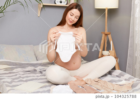 Smiling winsome pregnant brown haired caucasian woman sitting in bed with kids clothing planning packing things for maternity home. 104325788