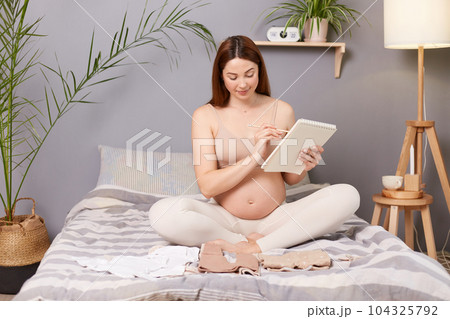 Smiling happy pregnant brown haired caucasian woman sitting in bed expecting baby holding paper notebook planning preparing for giving birth. 104325792