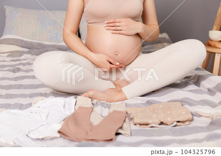 Unknown pregnant woman wearing beige top and leggins sitting in bed surrounded with tiny kid's clothing preparing for giving birth. Unknown pregnant woman wearing beige top and leggins sitting in bed surrounded with tiny kid's clothing preparing for giving birth. 104325796