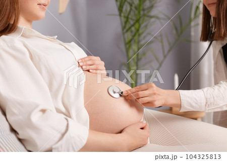 Unrecognizable pregnant female with bare belly and stethoscope doctor listening to baby heartbeat woman wearing white shirt appointment in hospital. 104325813