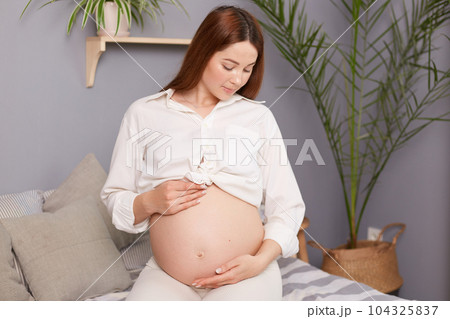 Beautiful expectant mother sitting on bed, touching her bare tummy with affection, happily waiting for her baby's arrival, wearing white shirt looking at her belly. Beautiful expectant mother sitting on bed, touching her bare tummy with affection, happily waiting for her baby's arrival, wearing white shirt looking at her belly. 104325837